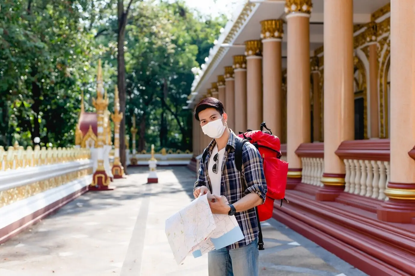 Ein junger asiatischer Rucksacktourist mit Gesichtsmaske steht an einem schönen thailändischen Tempel, hält eine Papierkarte in der Hand, prüft die Richtung und zeigt auf die Karte.